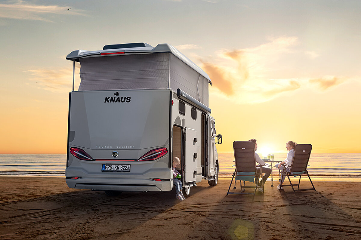 Ein Paar sitzt am Strand neben dem eigenen Wohnmobil und beobachtet den Sonnenuntergang.