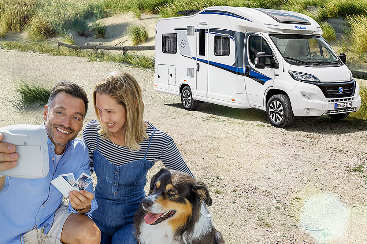 Ein Paar mit Hund macht ein Foto mit einer Sofortbildkamera vor dem gemieteten Wohnmobil.